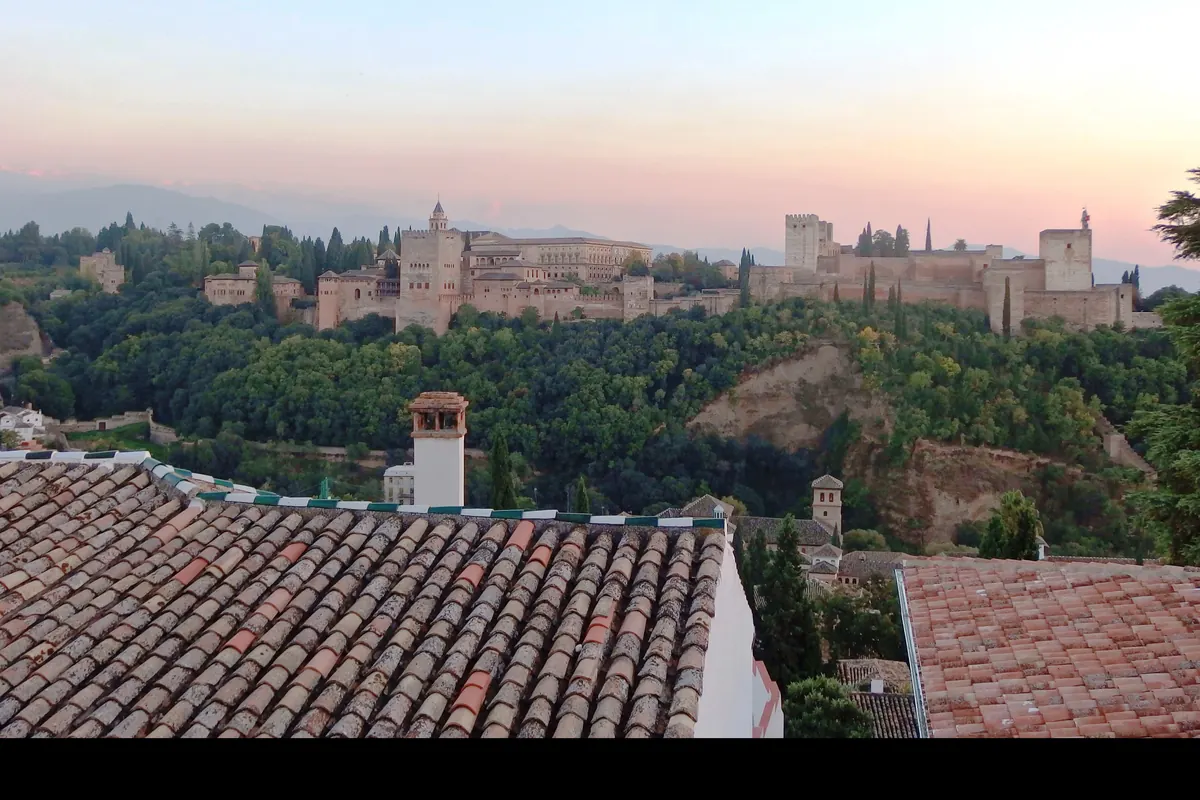 The Alhambra on its hill at dusk, seen across tiled rooftops from the Albaicín under a pink and blue sky