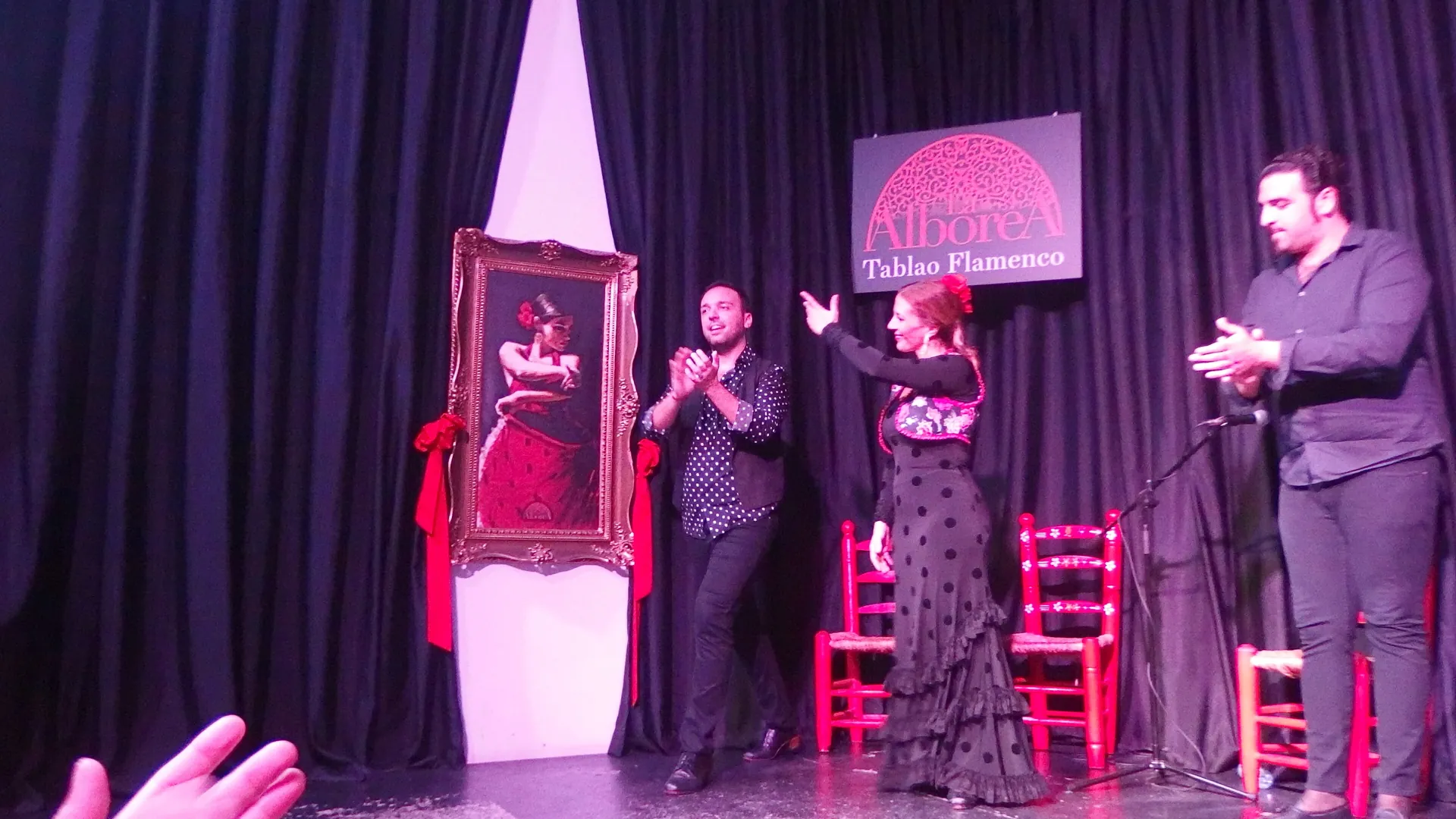Two flamenco performers clap and dance on the red-chaired stage of La Alboreá in Granada, under the lit venue sign and a framed painting of a dancer